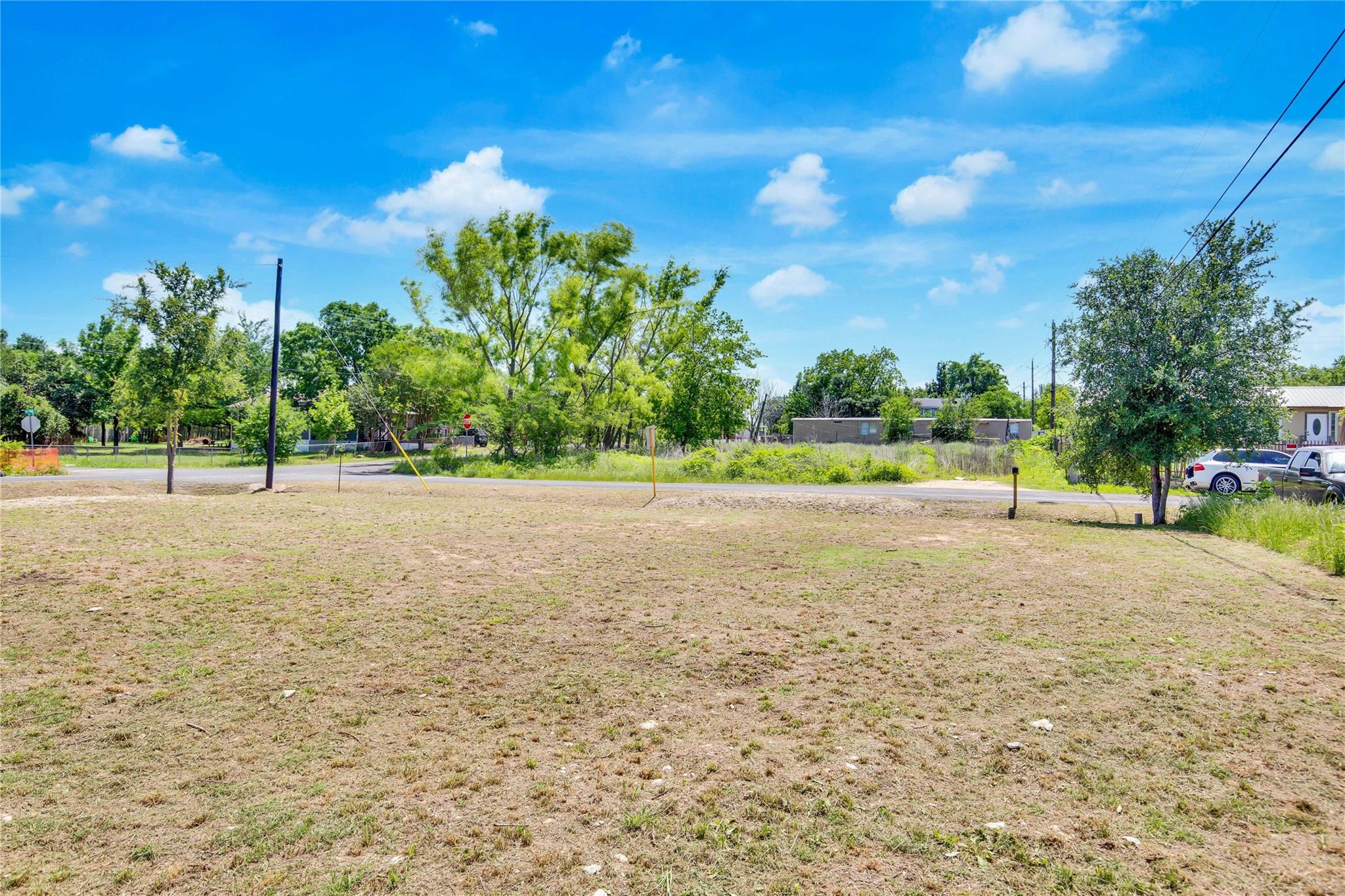 Vacant lot with dry grass, trees, and neighboring buildings across the road.