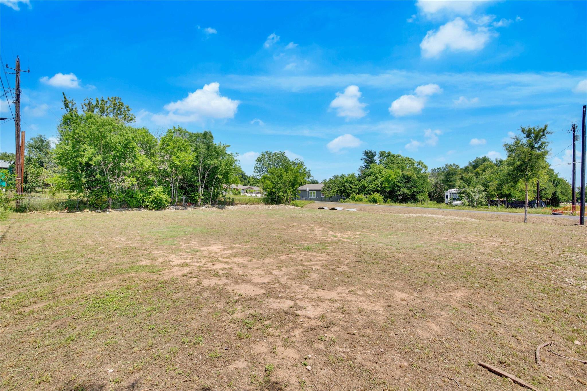 Vacant lot with patchy grass, mature trees along the edges, and neighboring homes in the distance.