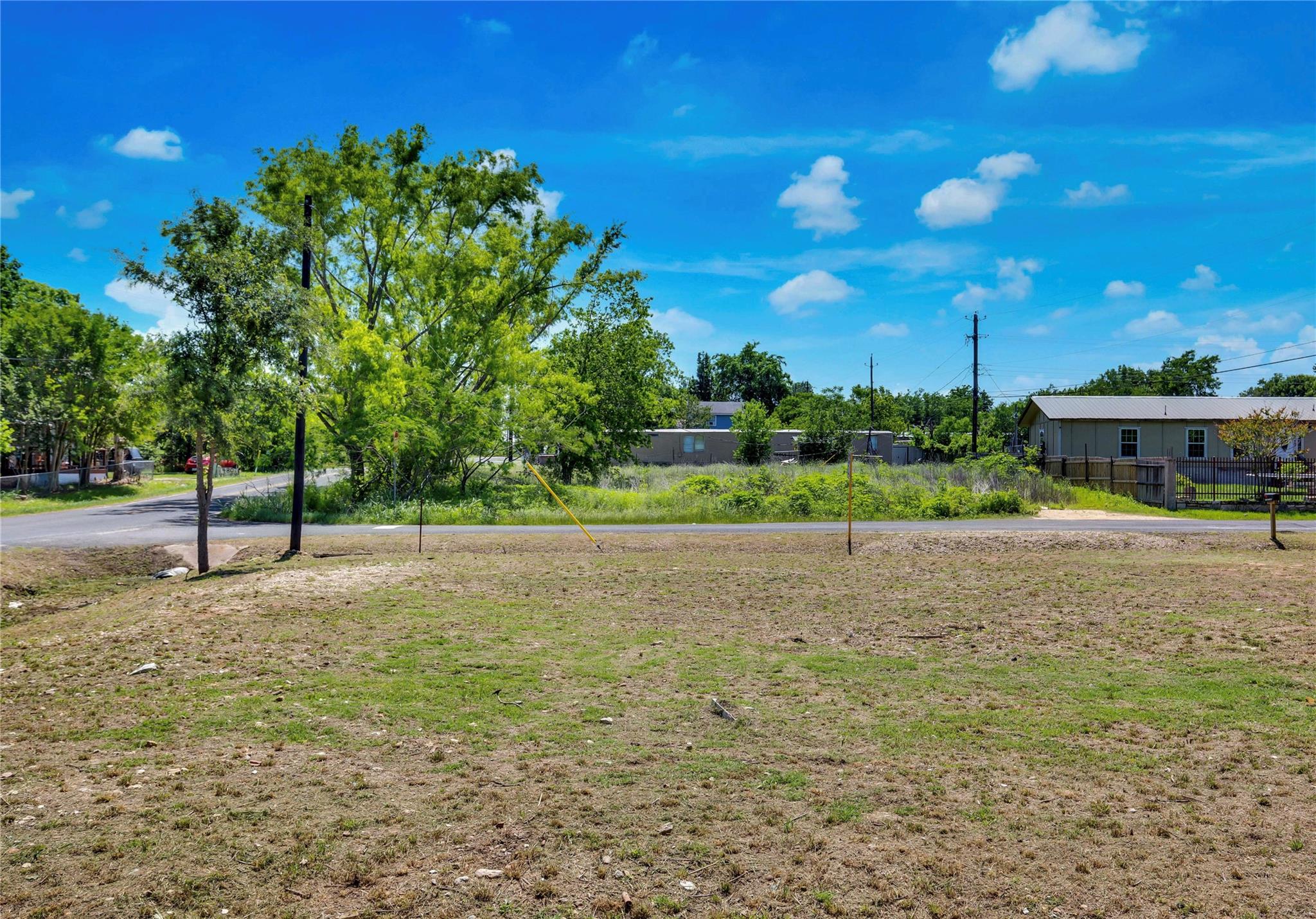 Vacant grassy lot beside a road, with mature trees and neighboring buildings across the street.