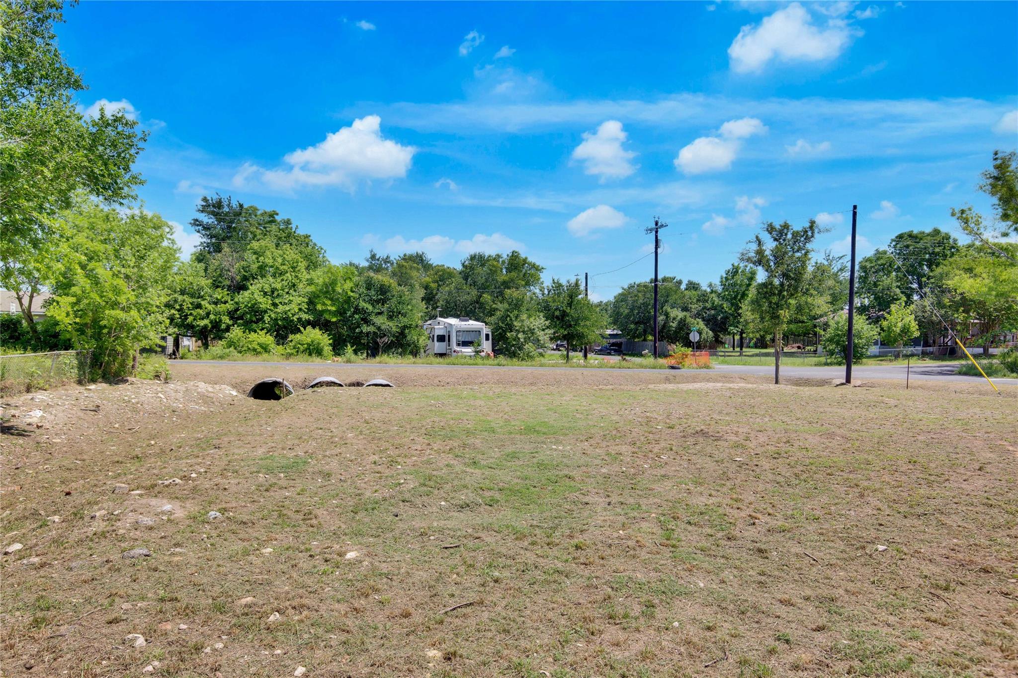 Mostly cleared grassy lot with roadside drainage culverts, trees, utility poles, and a recreational vehicle beyond the road.