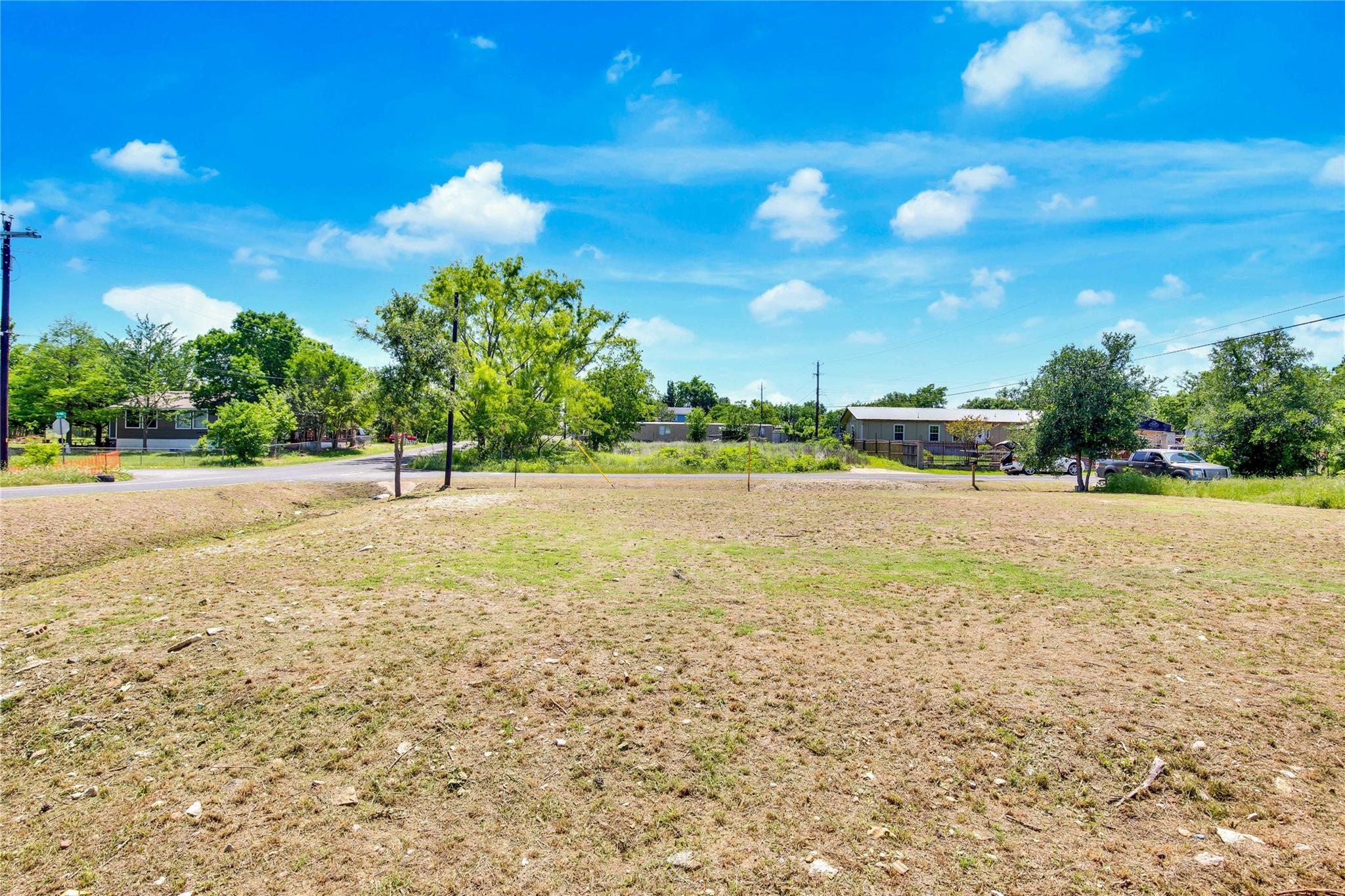 Vacant grassy lot beside a road, with scattered trees and neighboring homes.
