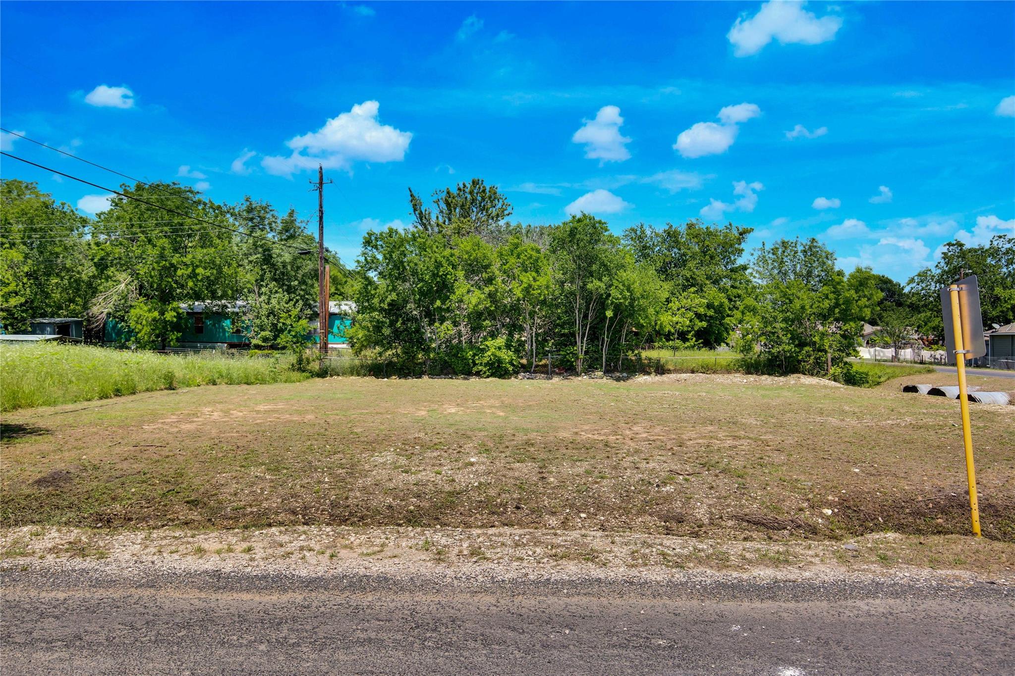 Vacant cleared lot with roadside frontage, sparse grass, and mature trees behind it.