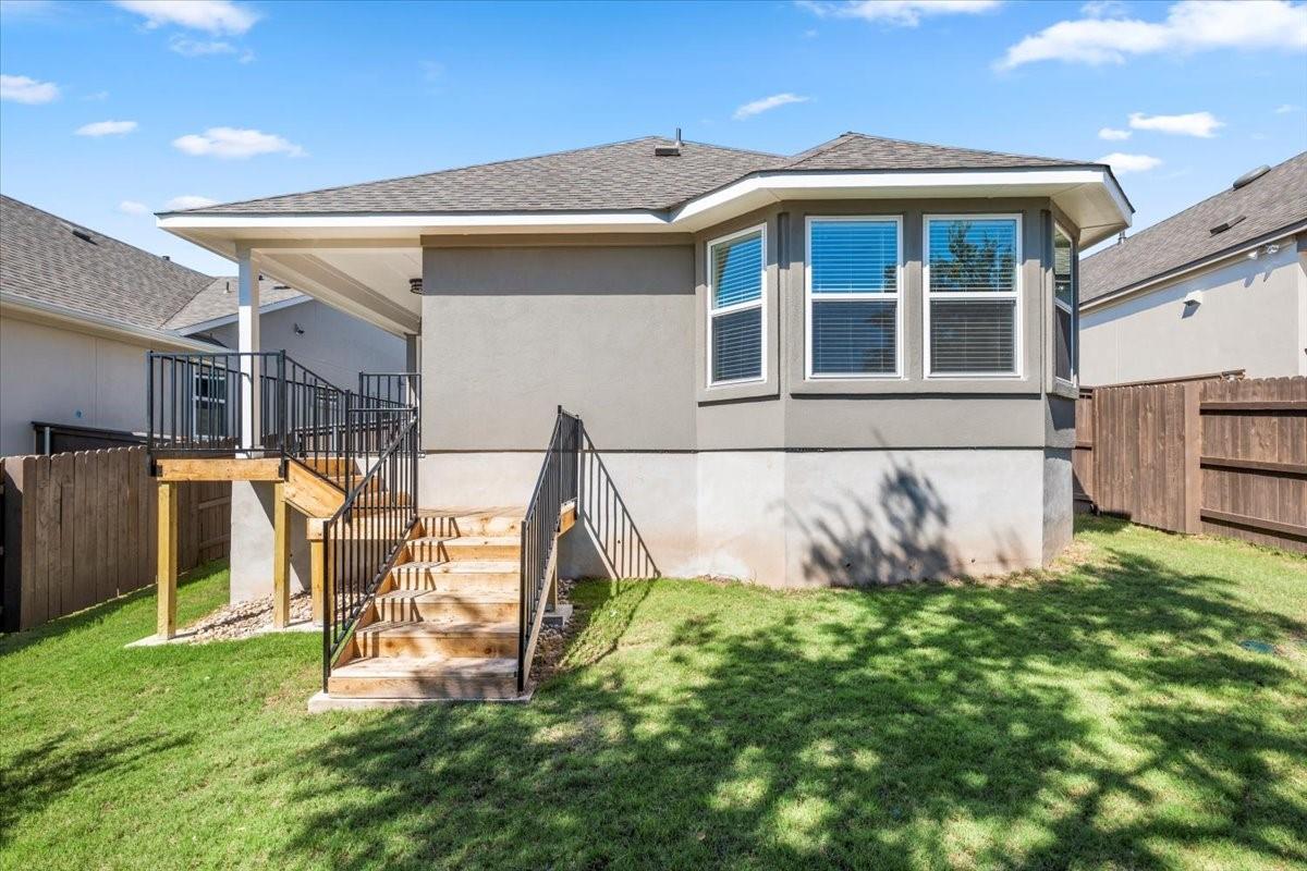 Rear of a single-story home with a covered raised deck, black-railed stairs, and fenced grassy yard.