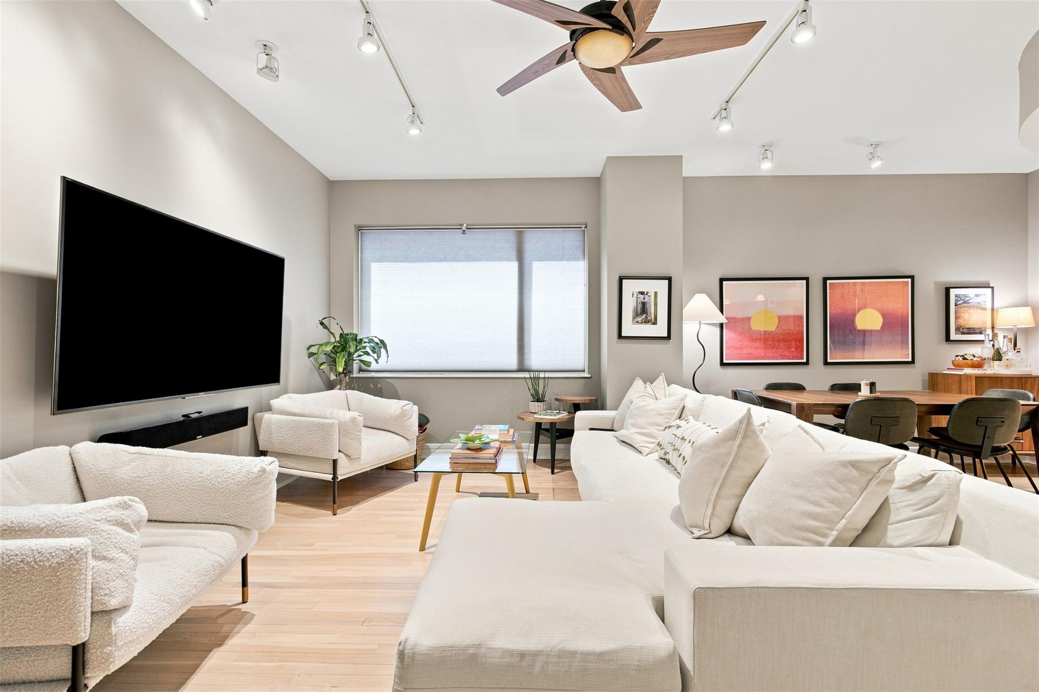 Furnished living room with white sofas, a wall-mounted TV, light wood floors, and an adjoining dining area.