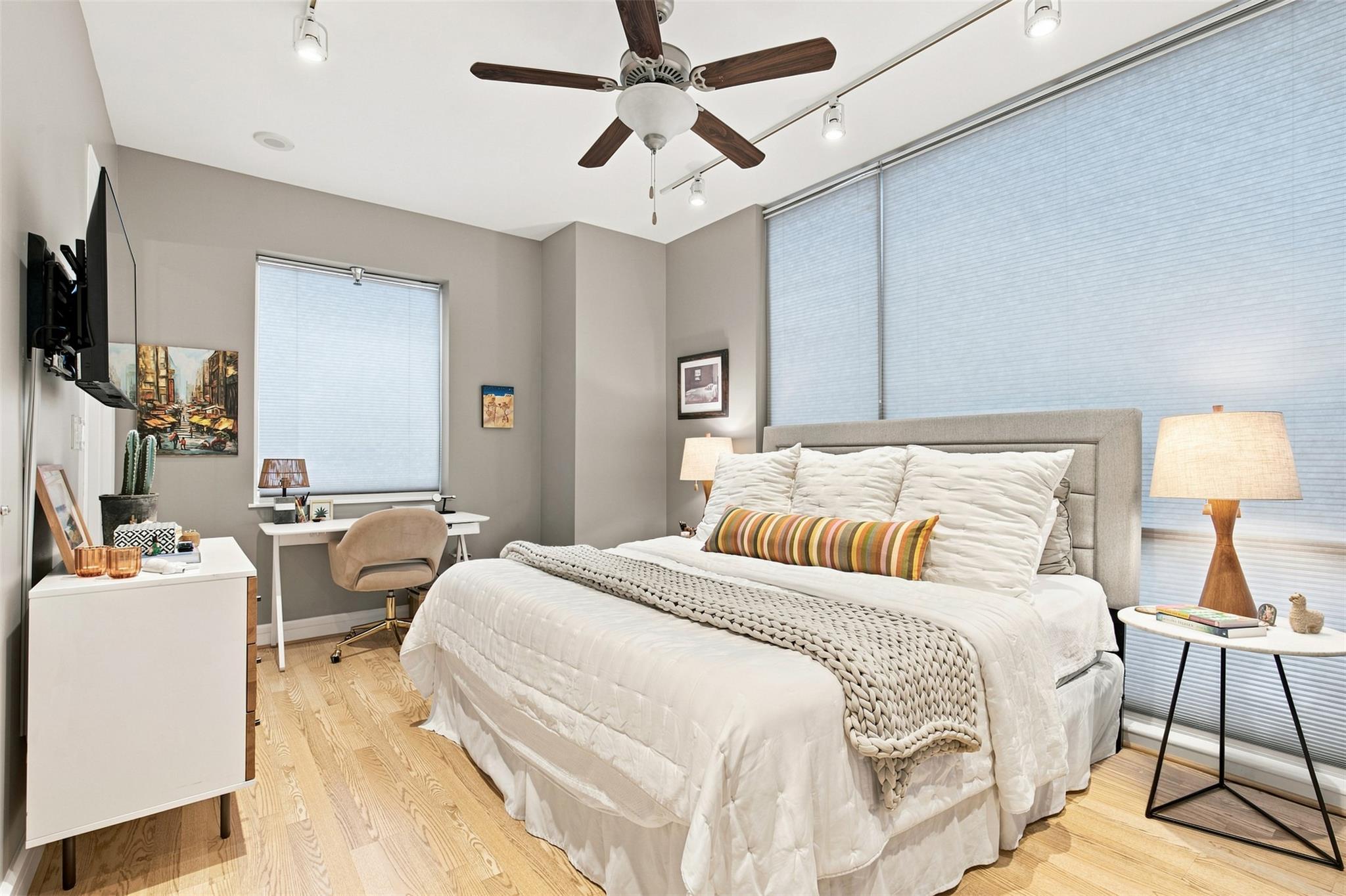 Furnished bedroom with a white bed, striped accent pillow, tall shaded windows, and pale wood floors.