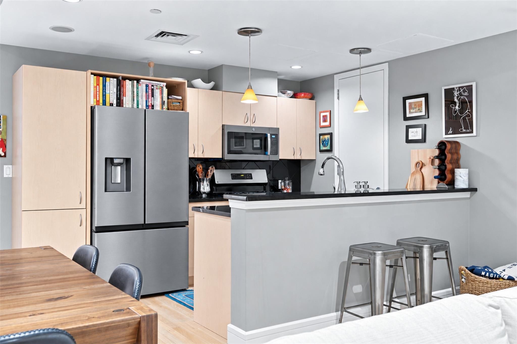 Modern kitchen with pale wood cabinets, stainless appliances, and a breakfast bar with metal stools.