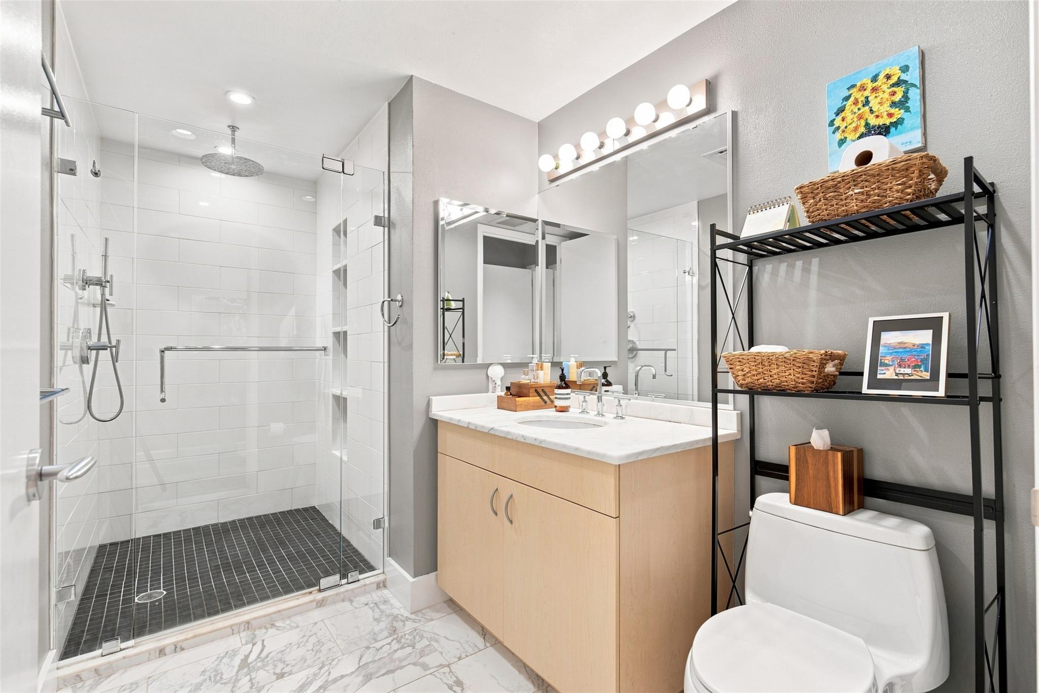 Bathroom with a glass walk-in shower, light wood vanity, and black shelving beside the toilet.