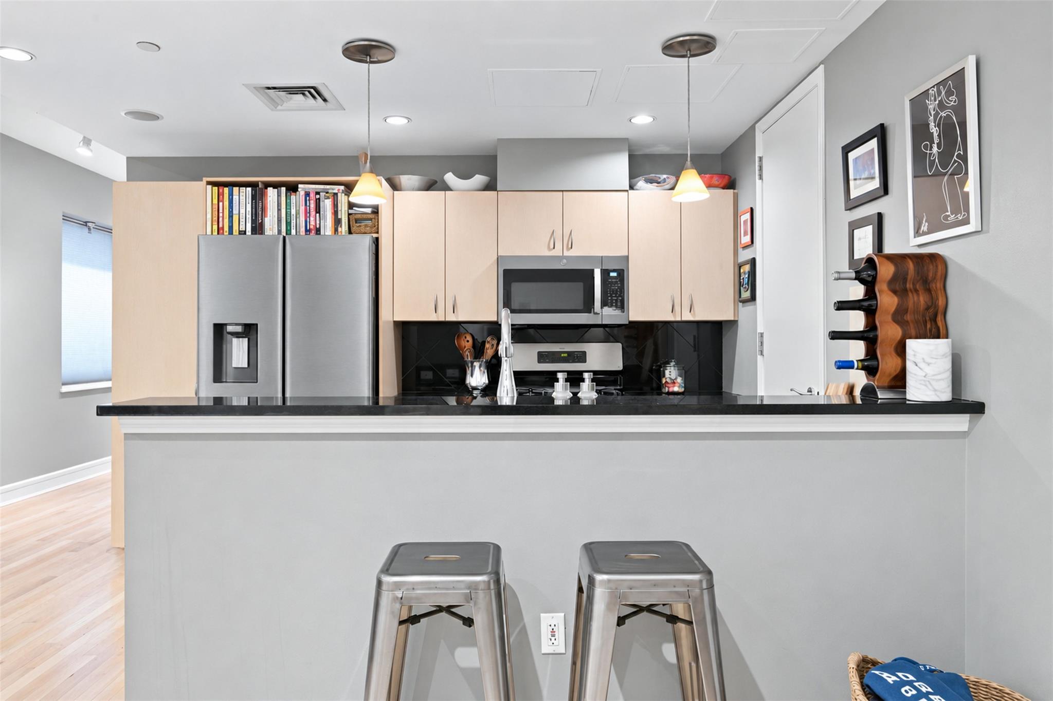 Condo kitchen with pale wood cabinets, stainless steel appliances, and two stools at a black breakfast bar.