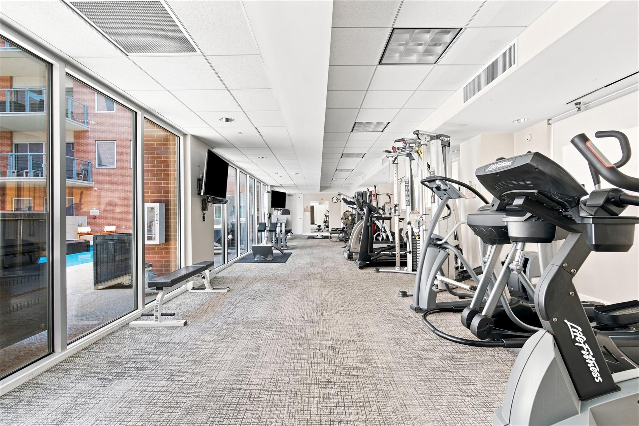 Fitness center with cardio and weight machines beside floor-to-ceiling windows overlooking a pool.