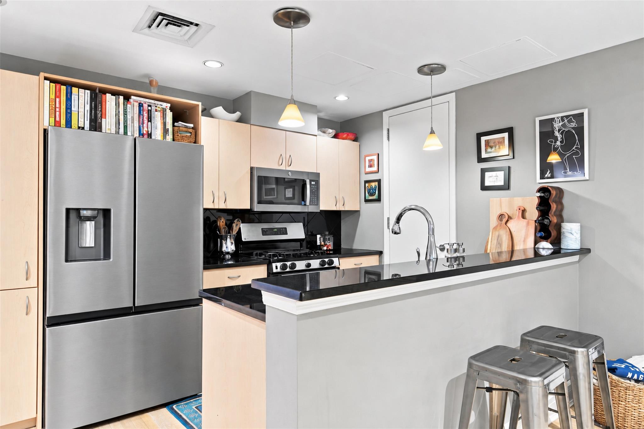 Kitchen with light wood cabinets, stainless steel appliances, black counters, and a breakfast bar with metal stools.