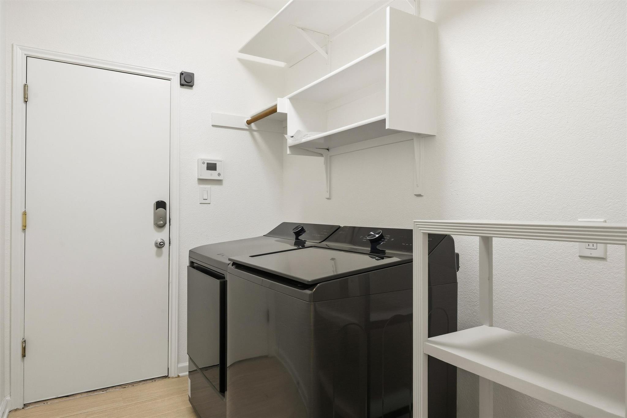 Laundry room with dark washer and dryer beneath white wall-mounted shelves.