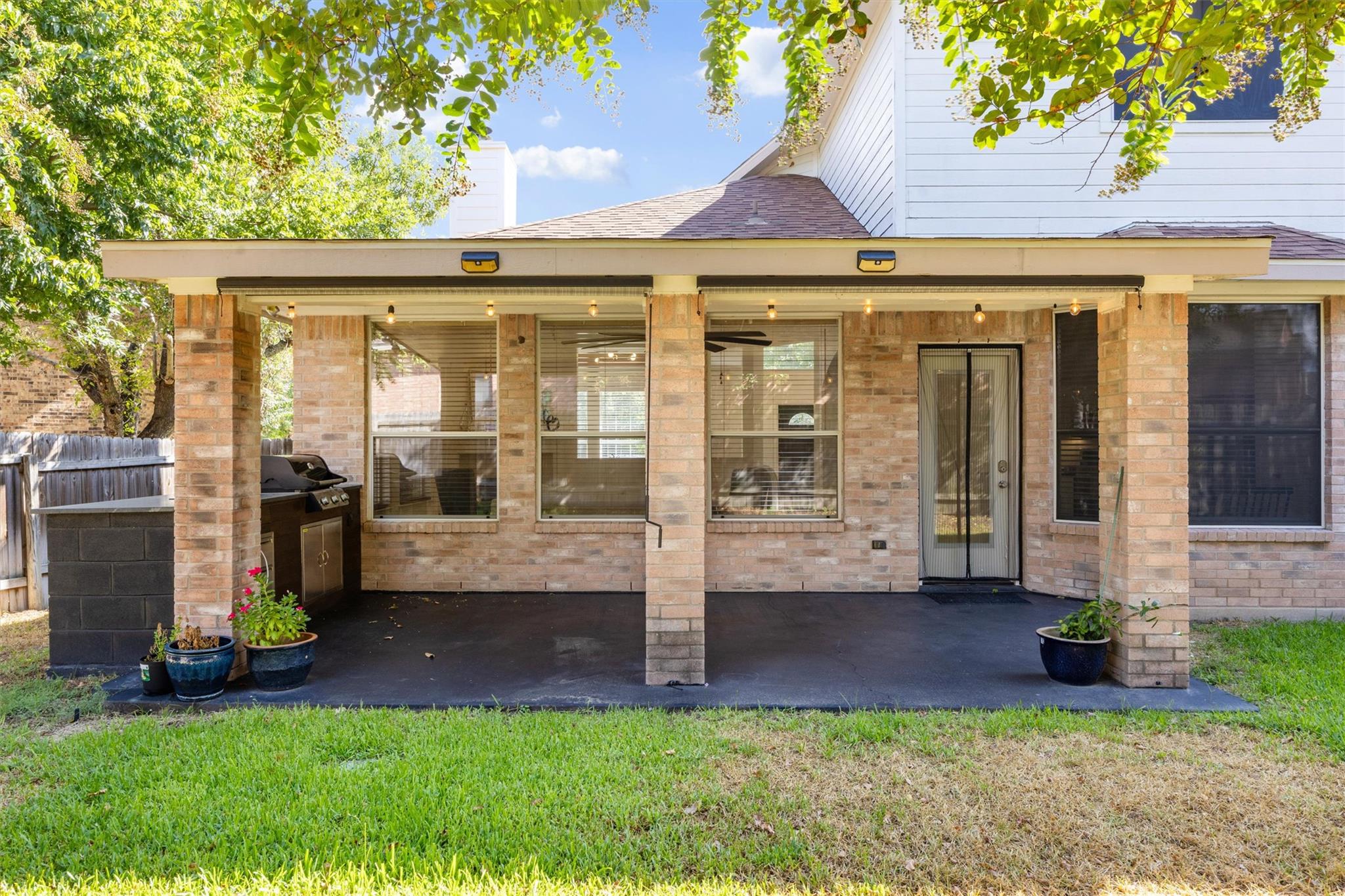 Brick home’s covered back patio with string lights, a grill, and potted plants.