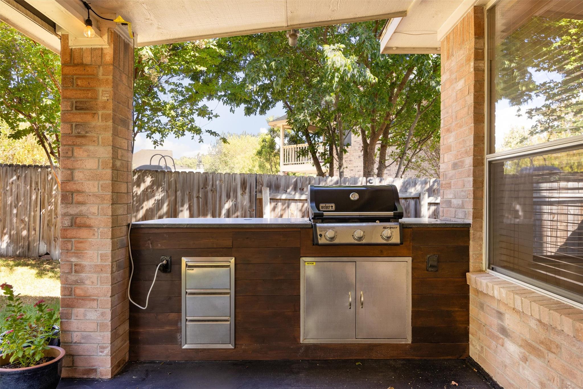 Covered patio outdoor kitchen with built-in grill, stainless cabinets, and a fenced leafy yard.