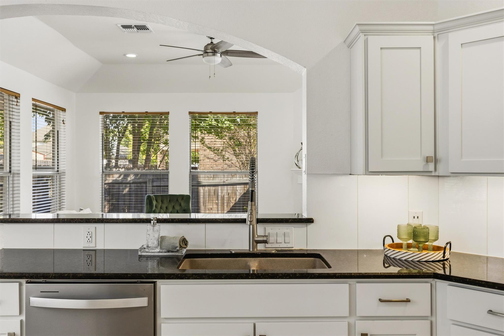Bright kitchen with white cabinets, dark counters, a stainless-steel sink, and windows overlooking a fenced yard.