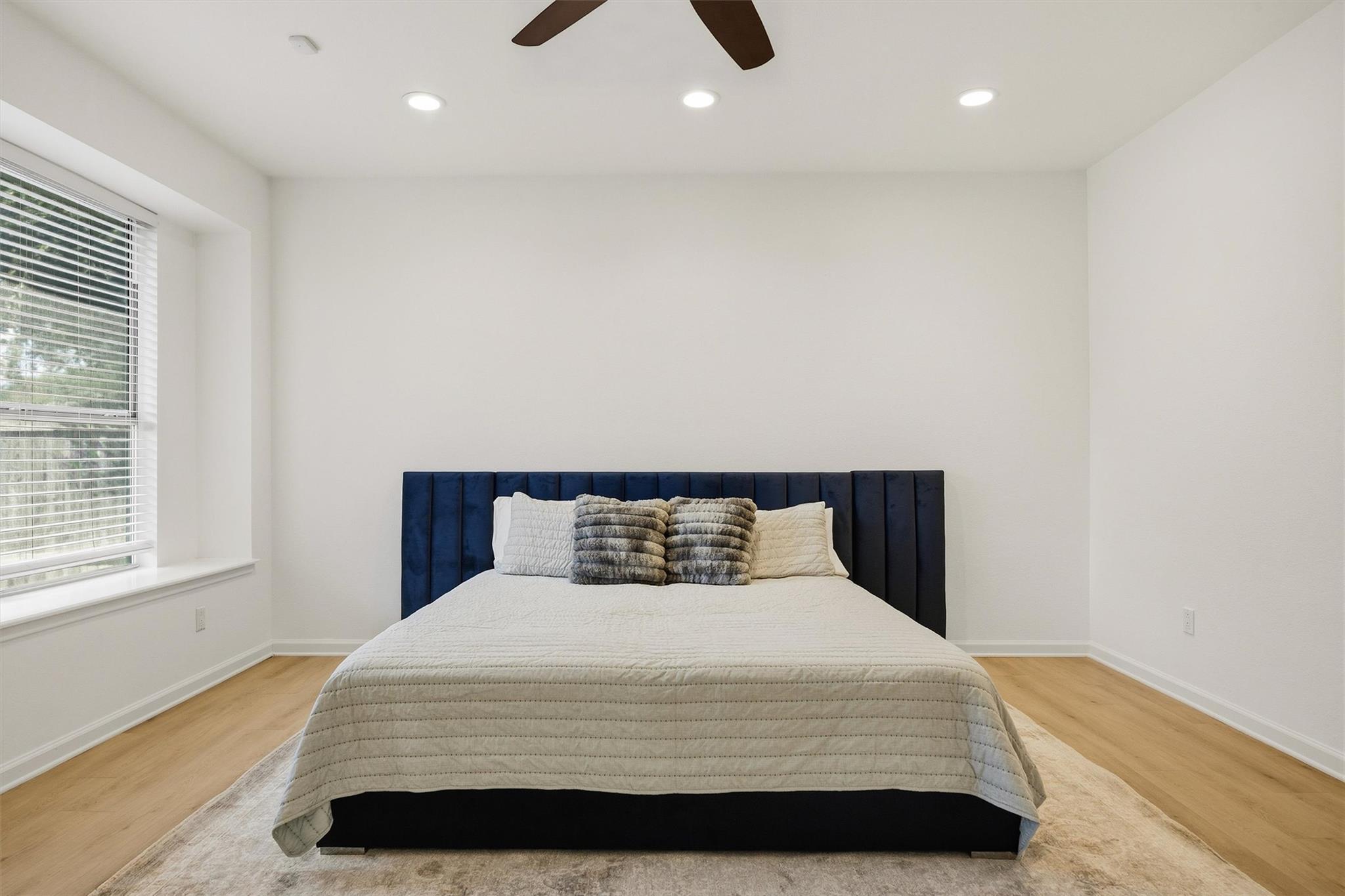 Bedroom with a white-quilted bed, navy upholstered headboard, and large window with blinds.