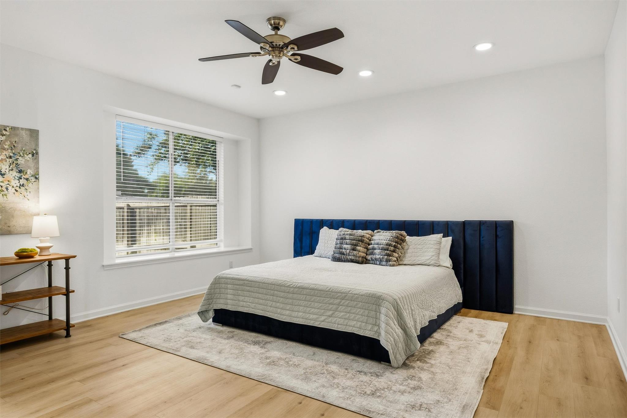 Furnished bedroom with a dark blue headboard, large window, ceiling fan, and light wood flooring.