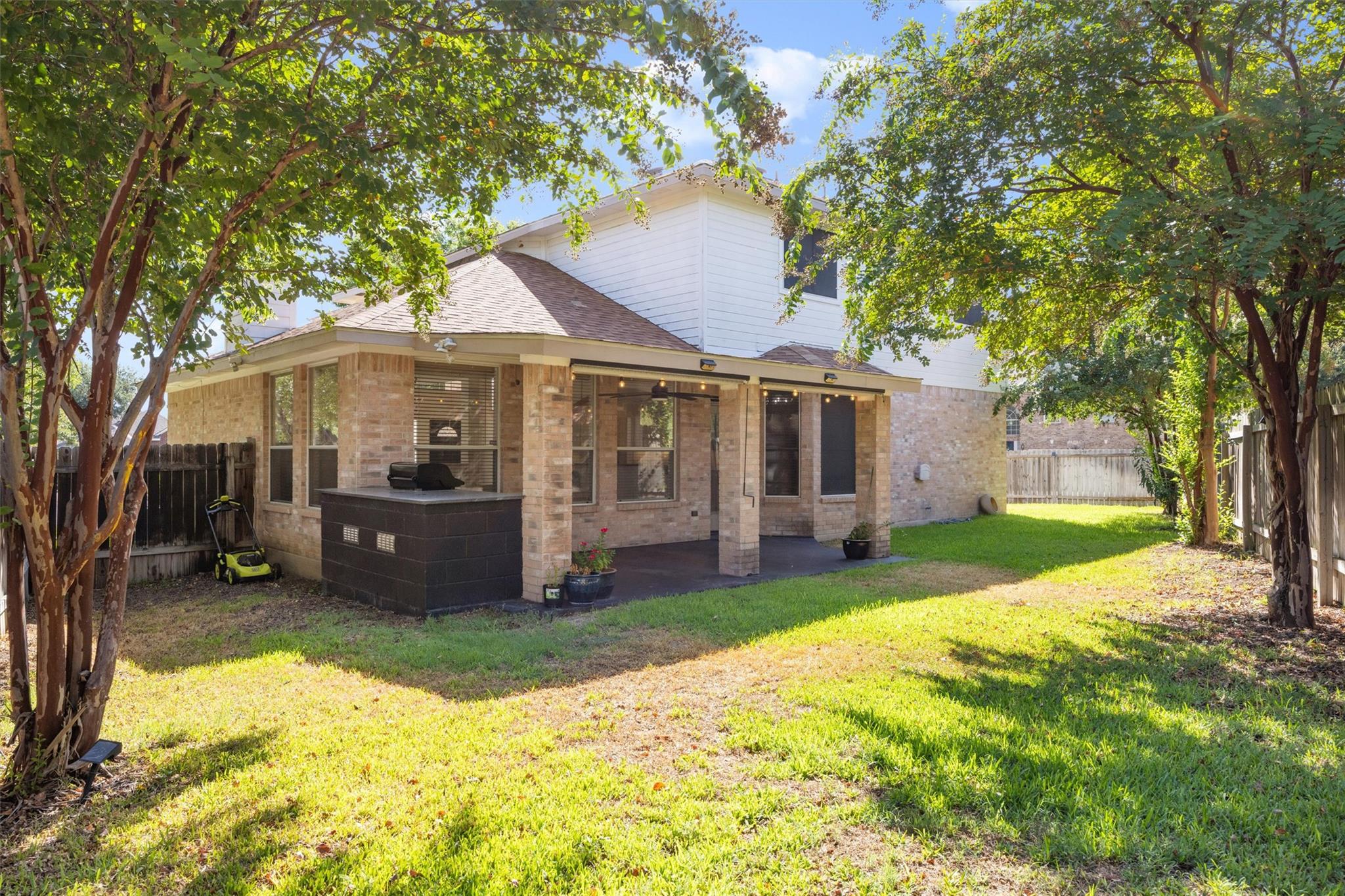 Rear of a brick house with a covered patio and grill beneath mature trees.