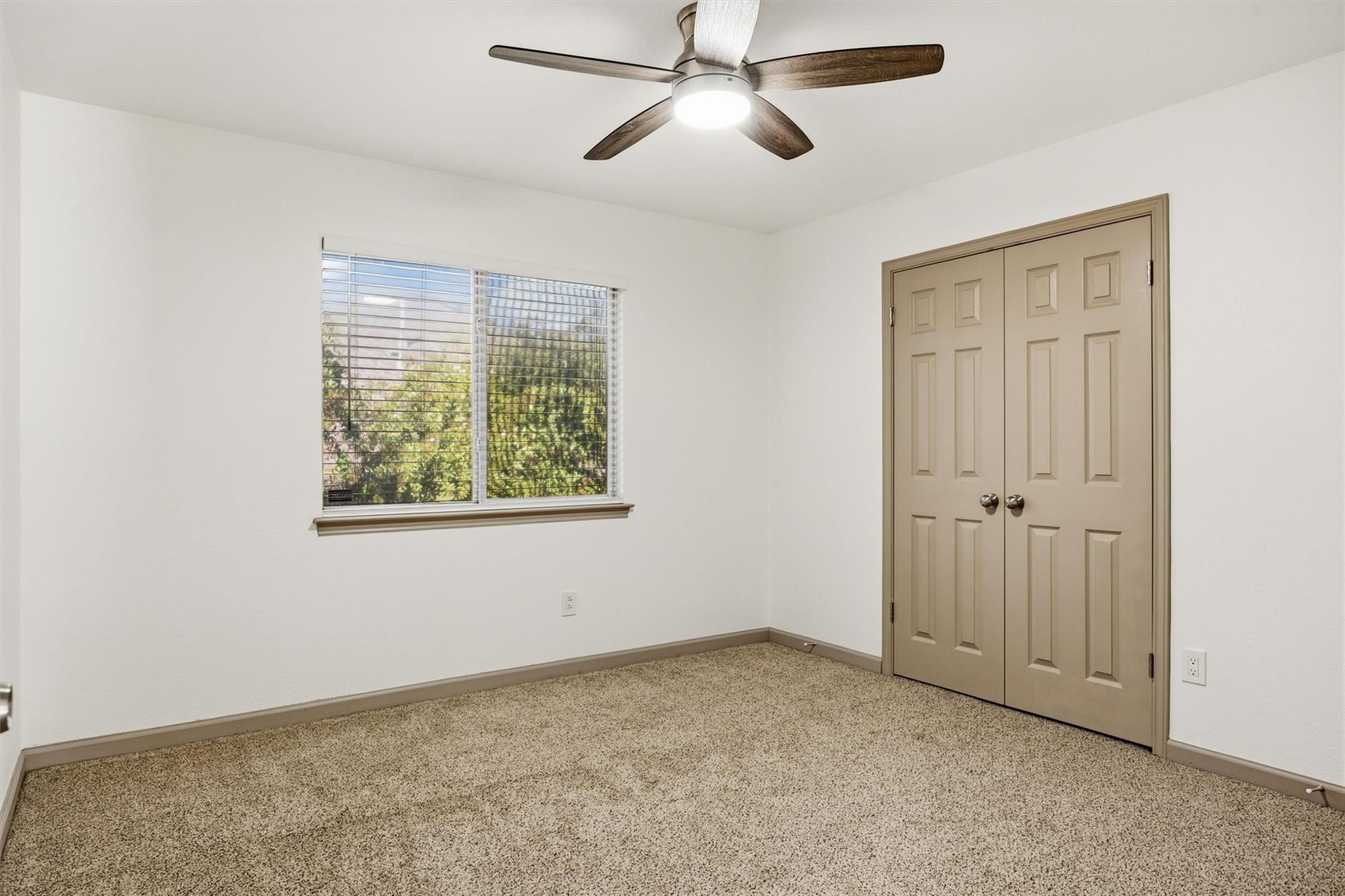 Empty bedroom with beige carpet, double closet doors, a window with blinds, and a ceiling fan.