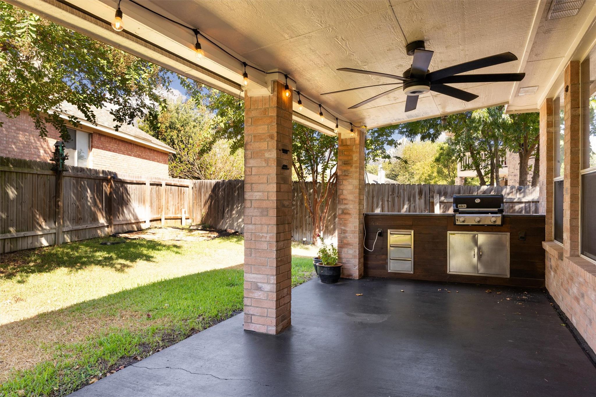 Covered patio with a built-in grill, ceiling fan, string lights, and fenced grassy yard.