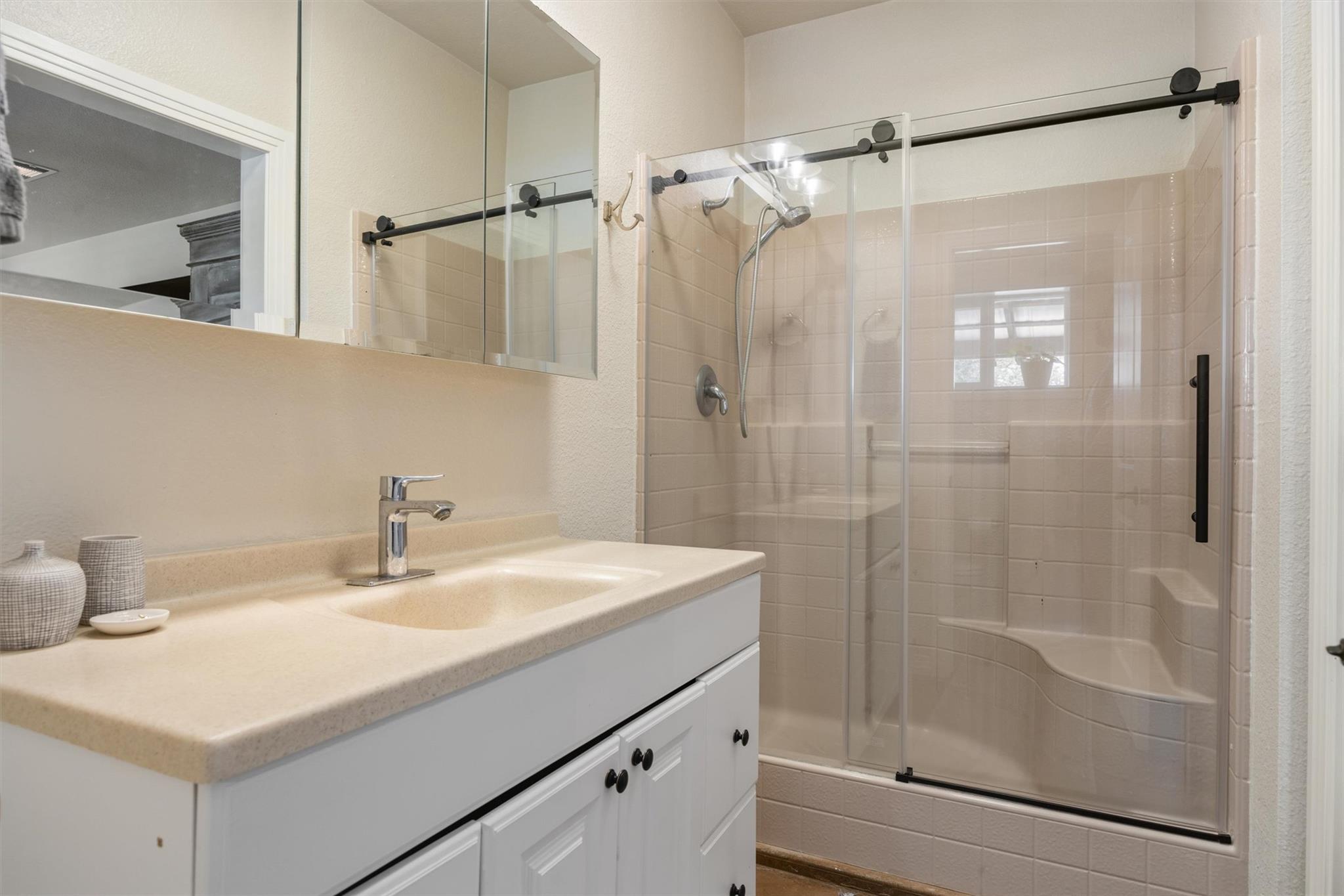 Bathroom with a white vanity and beige countertop beside a glass-enclosed tiled shower with a built-in bench.