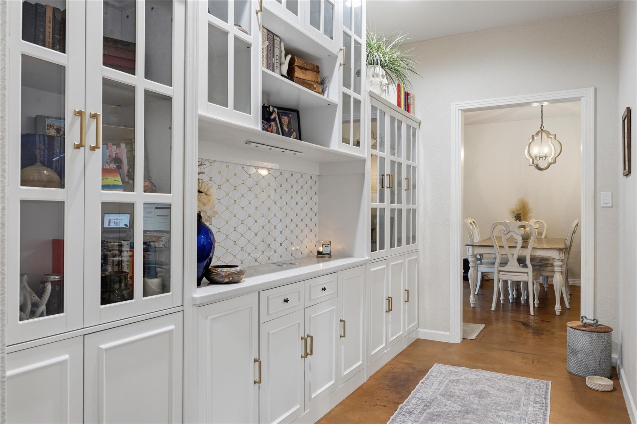 White built-in cabinets with geometric tile backsplash beside a dining room with a chandelier.