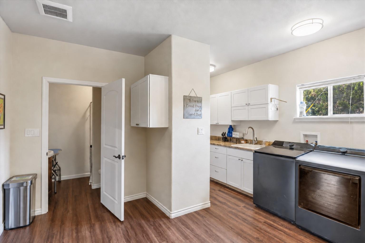 Laundry room with white cabinets and sink, two dark machines, and an open doorway.