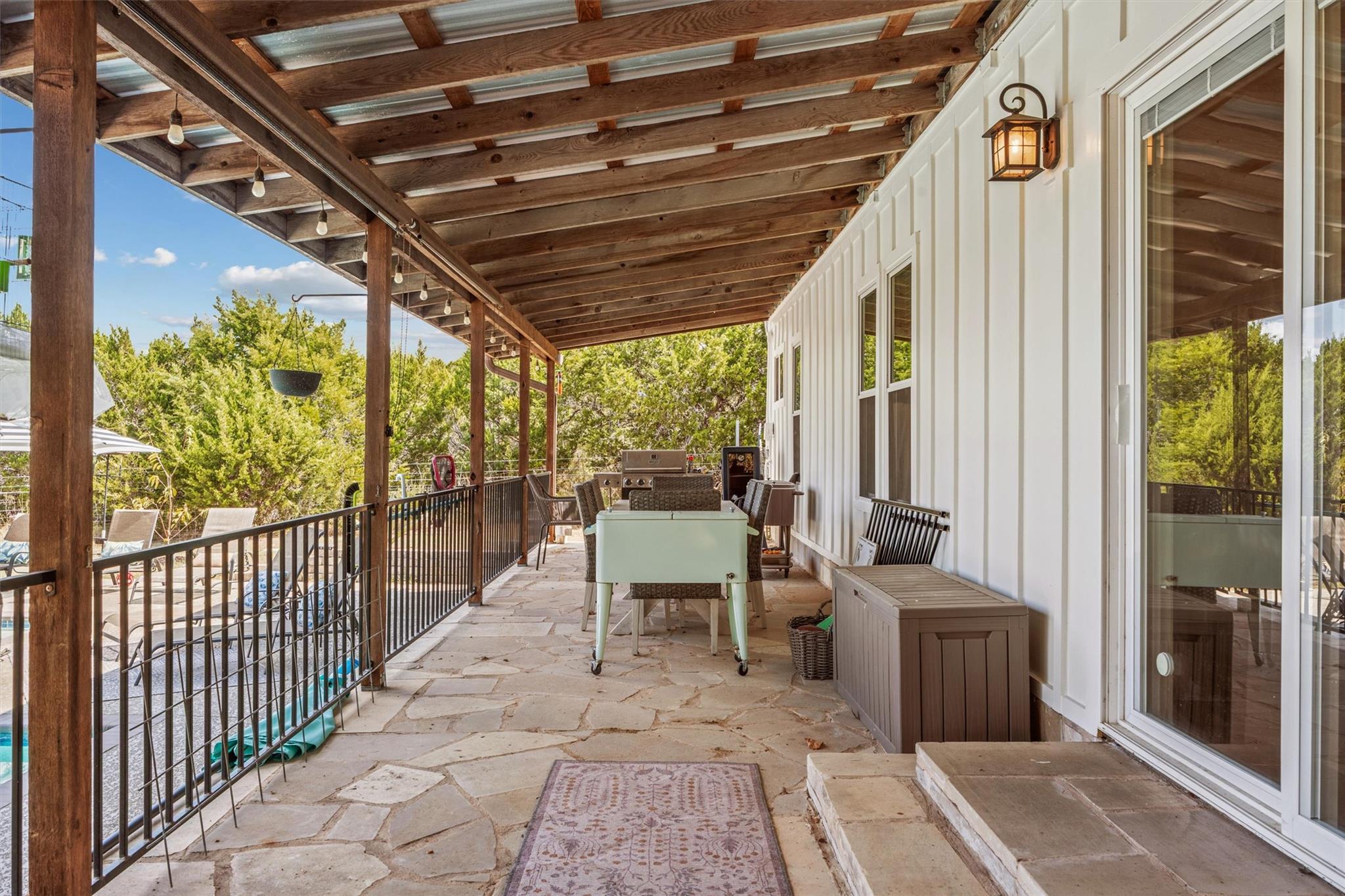 Covered stone patio with timber beams, outdoor dining furniture, and a swimming pool beyond the railing.
