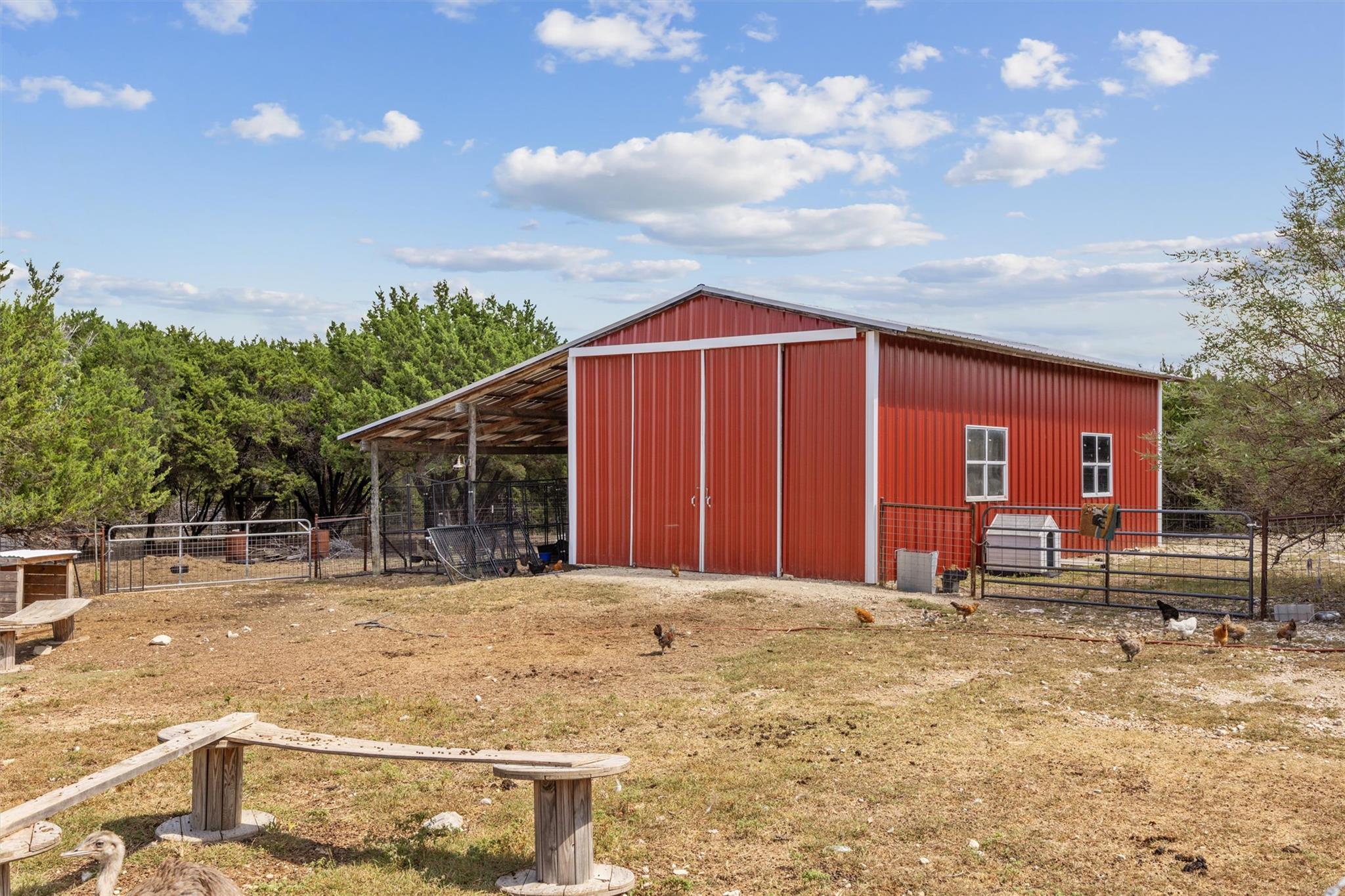 Red metal barn with a covered lean-to, fenced animal pens, and chickens in the yard.