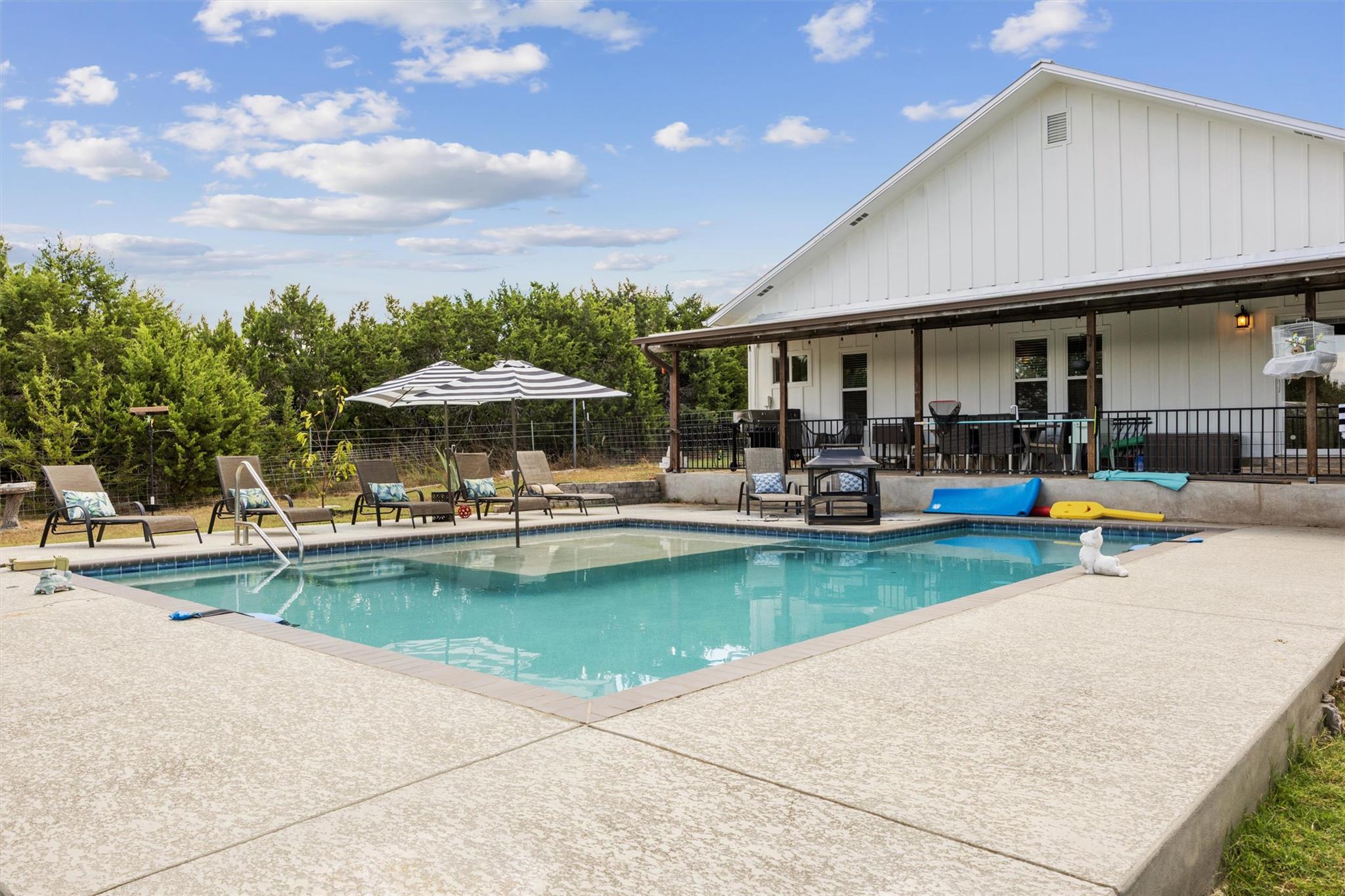 Backyard swimming pool beside a white house with a covered patio and striped umbrellas.