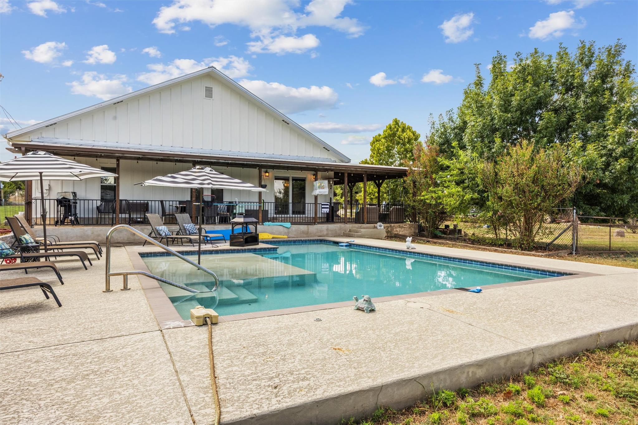 Swimming pool beside a white home, with a covered patio and striped umbrellas.