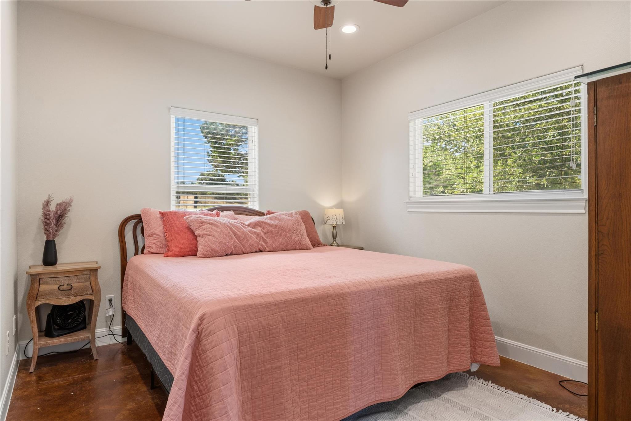 Bedroom with a dusty-pink quilted bed, white blinds, and polished brown flooring.