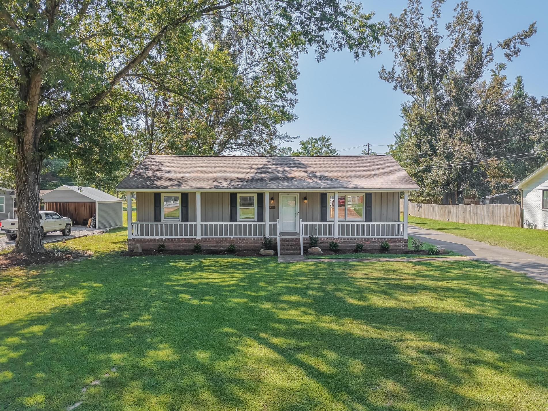 Gray single-story house with a long covered porch, brick foundation, and shaded front lawn.