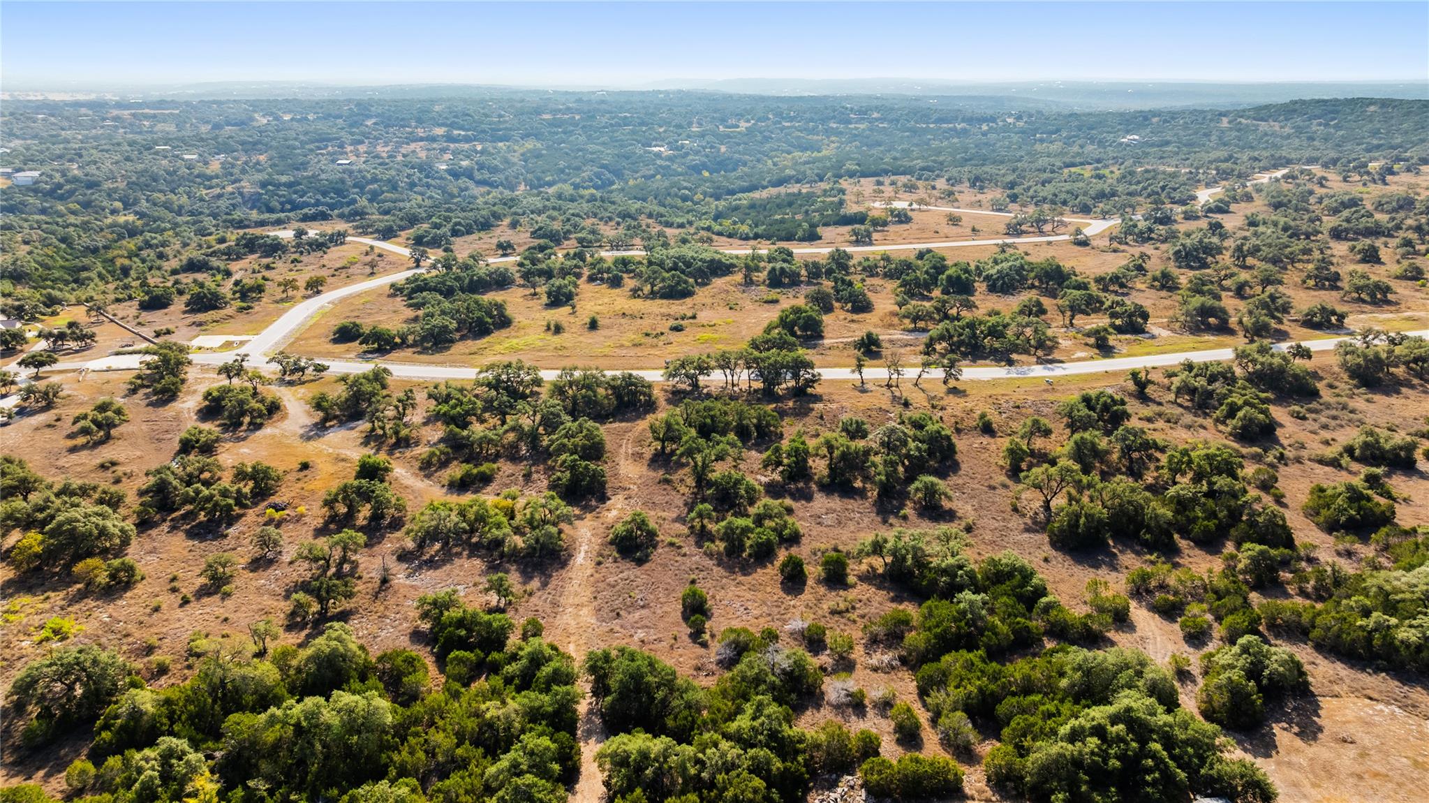 Aerial view of dry, tree-dotted land with winding roads and wooded hills in the distance.