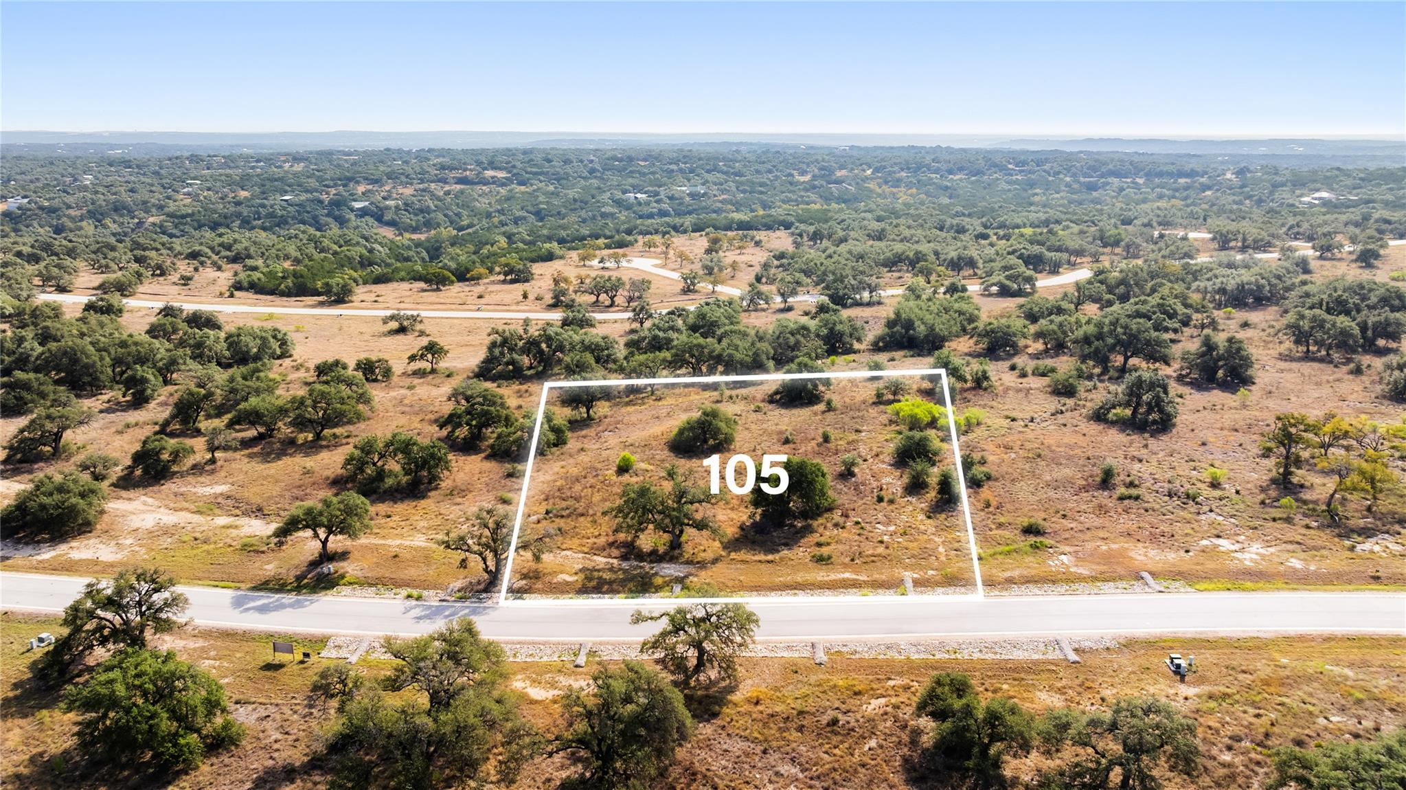 Vacant lot 105 outlined beside a paved road, surrounded by scattered trees and open terrain.