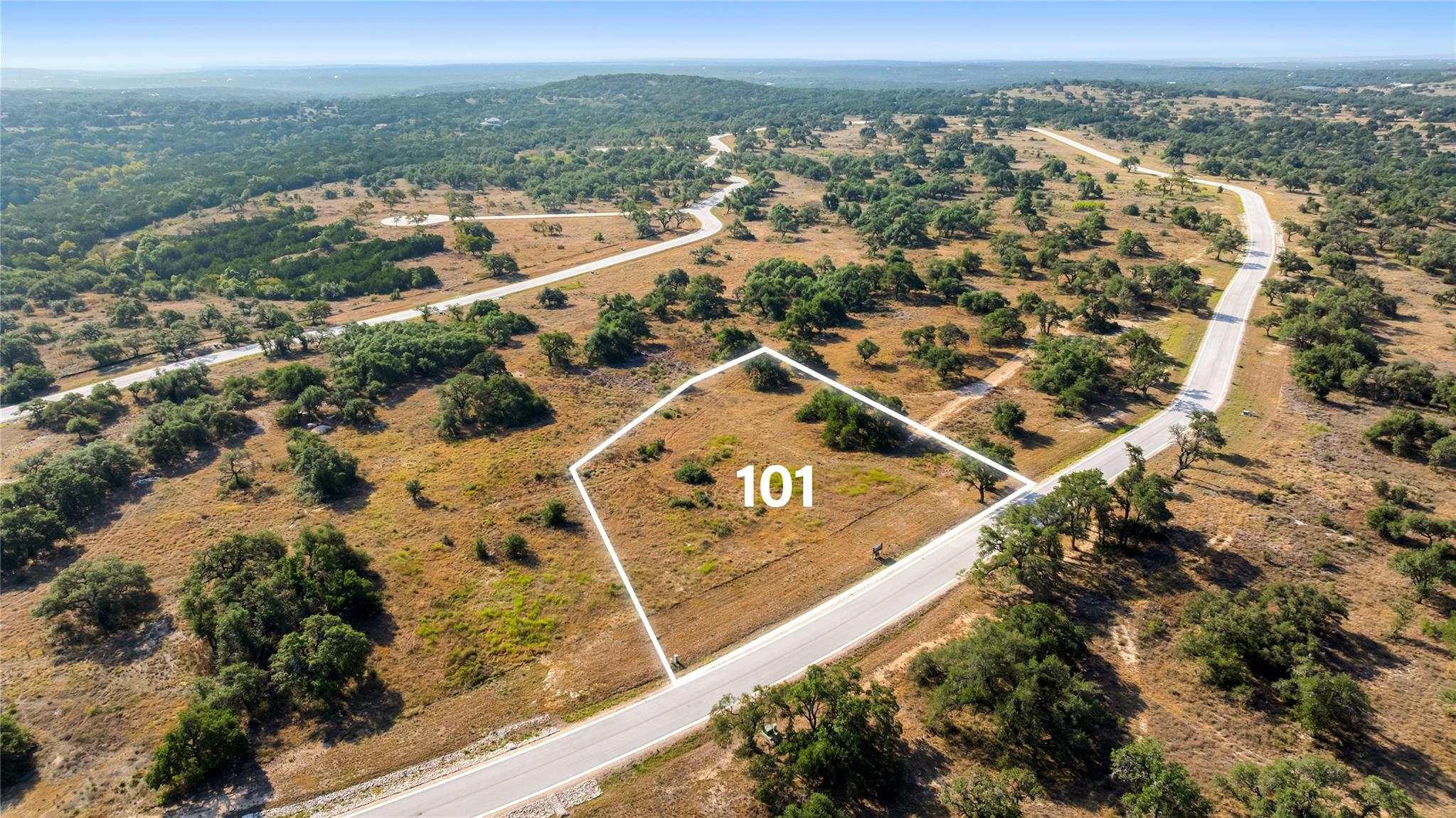 Aerial view of vacant Lot 101 outlined beside a paved road, surrounded by brush and scattered trees.
