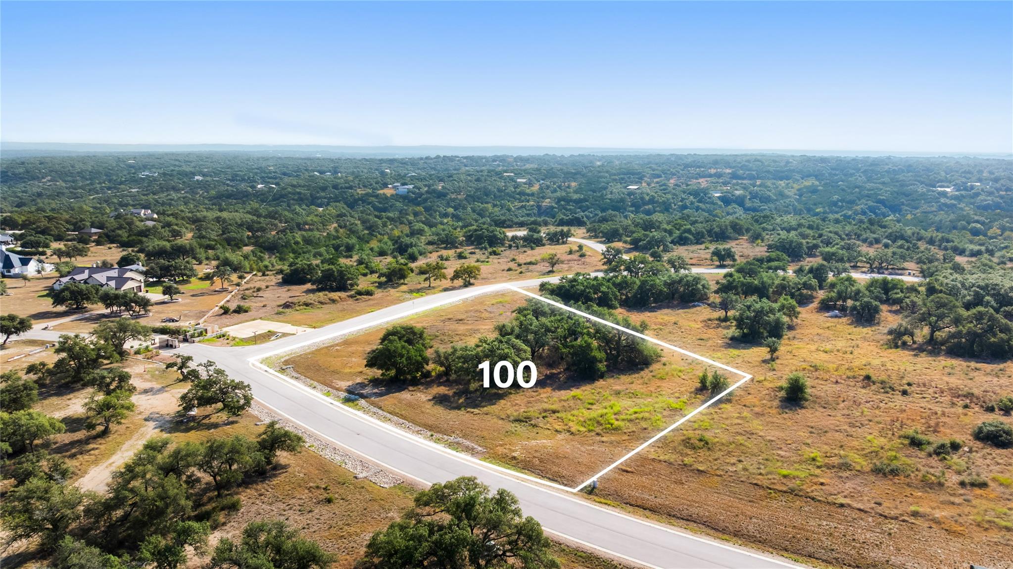 Aerial view of vacant Lot 100, outlined beside a paved road amid scattered trees and dry grass.