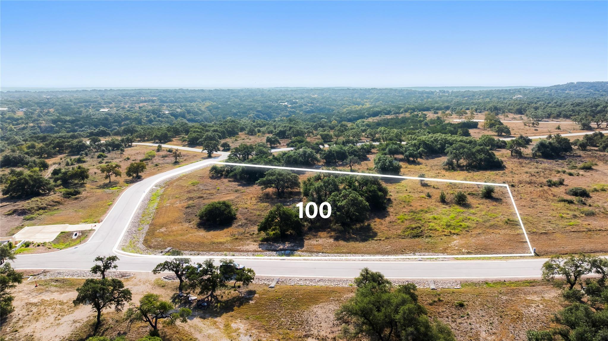 Aerial view of Lot 100 outlined beside paved roads, with scattered trees and wooded countryside beyond.