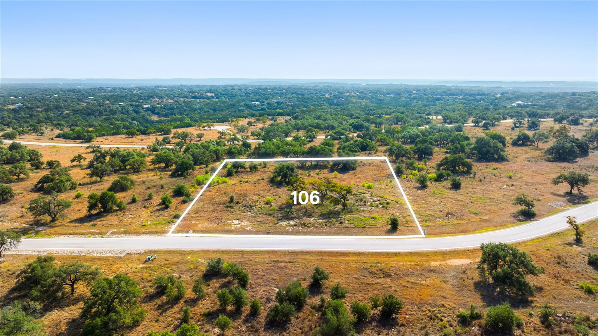 Vacant brush-covered lot 106 outlined beside a paved road, with scattered trees and wooded countryside beyond.