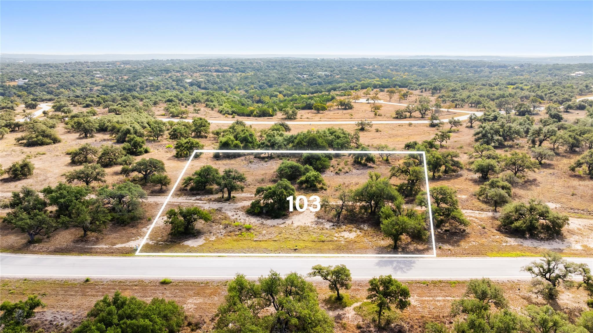 Aerial view of Lot 103, an oak-dotted vacant parcel outlined in white beside a paved road.