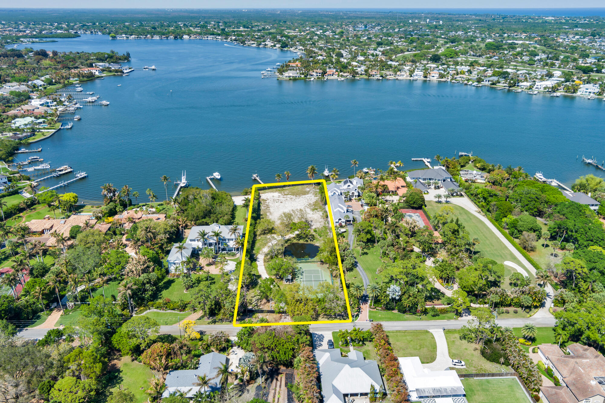 Aerial view of a waterfront property outlined in yellow, with a tennis court and pond.