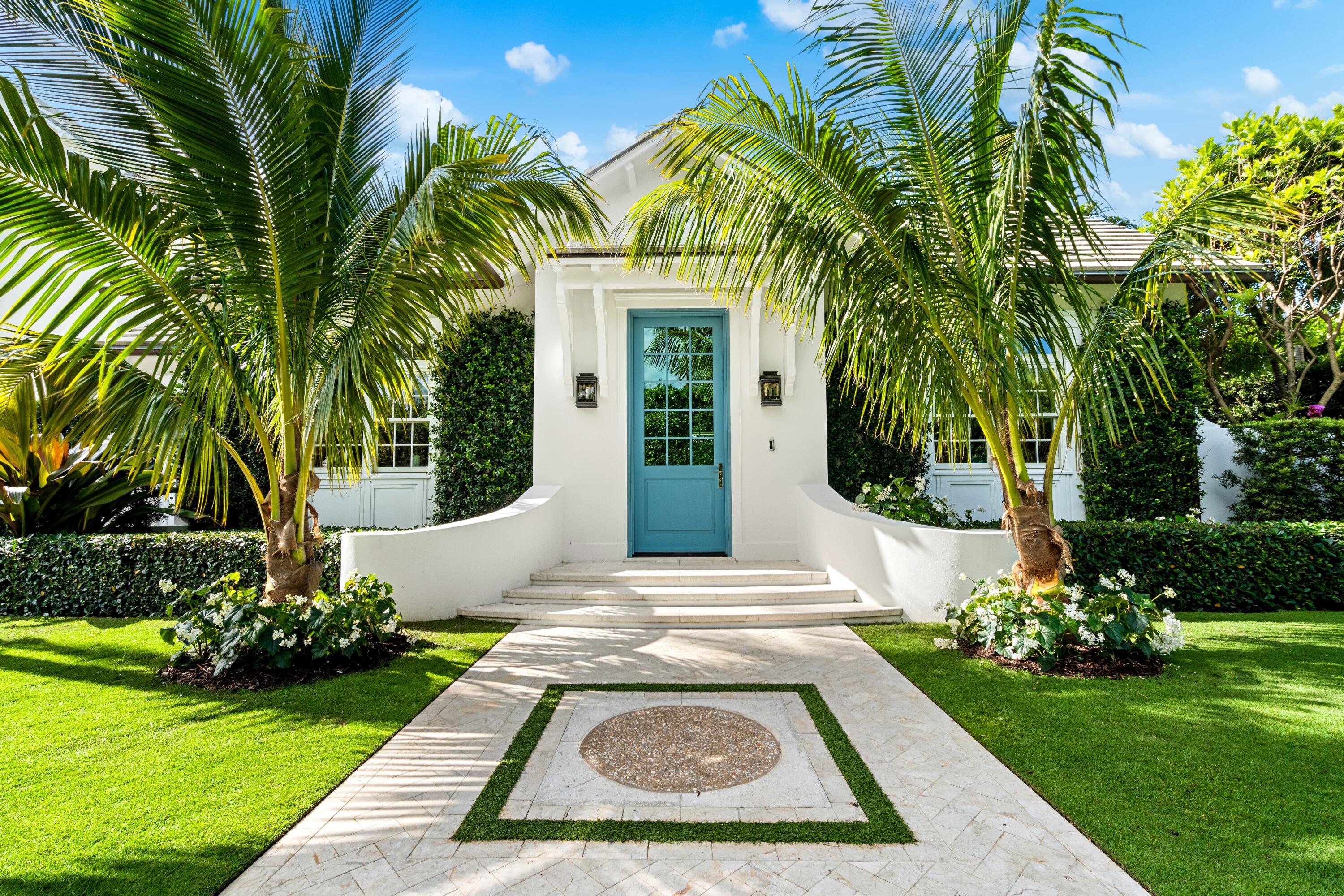 White home entrance with a blue door, curved stone steps, and palm trees framing the walkway.