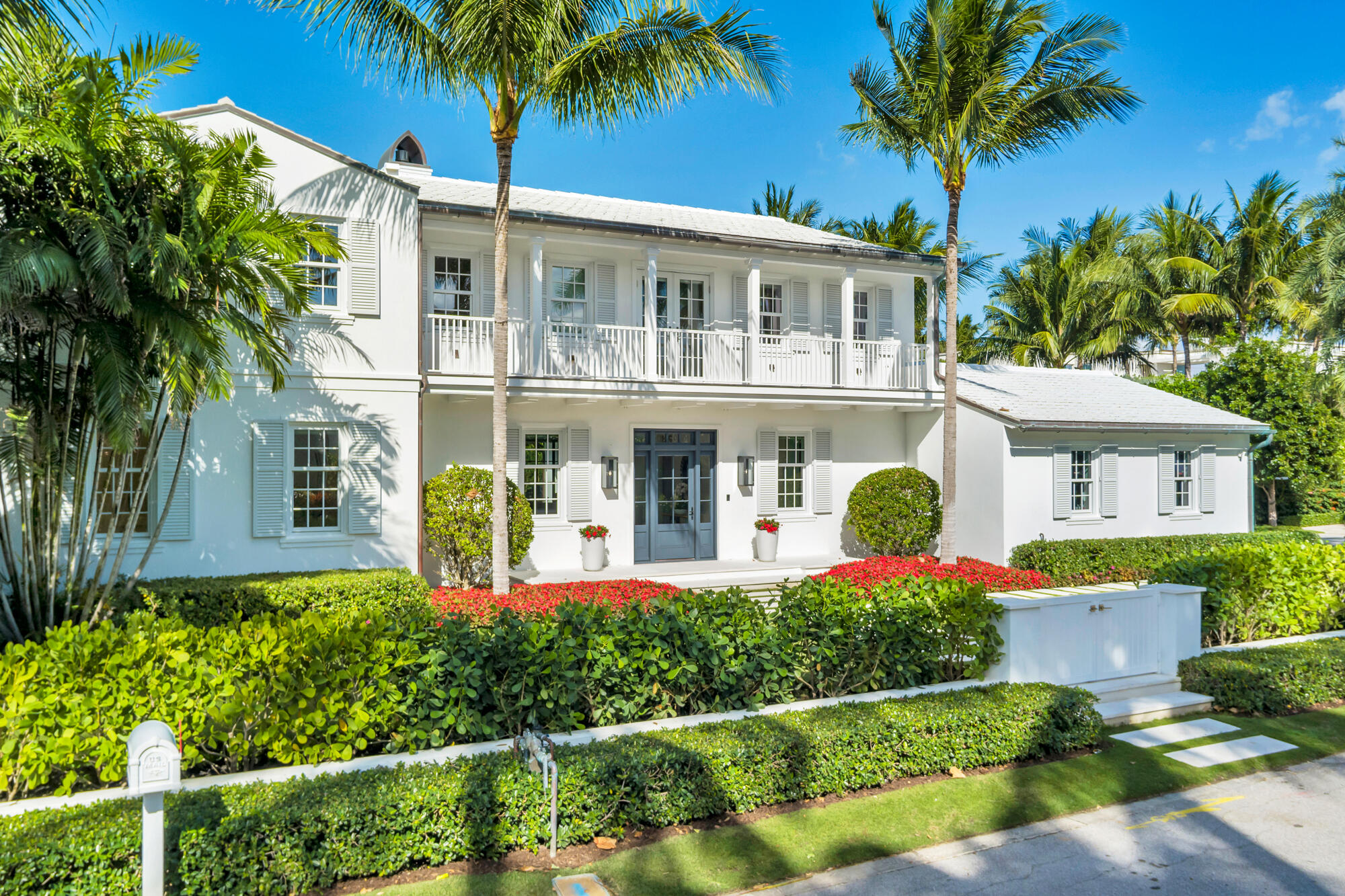 White two-story home with a long balcony, palm trees, and red flowers along the front hedge.