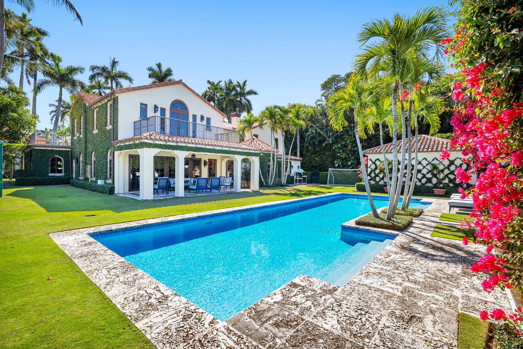 Mediterranean-style home behind a turquoise pool, with a covered patio, palms, and vivid bougainvillea.