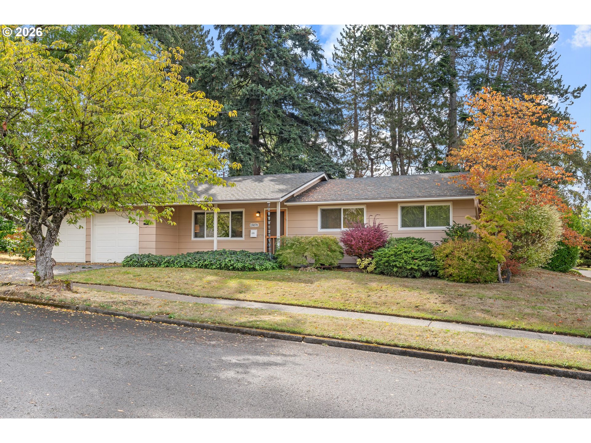 Single-story tan house with an attached garage, leafy landscaping, and autumn trees.