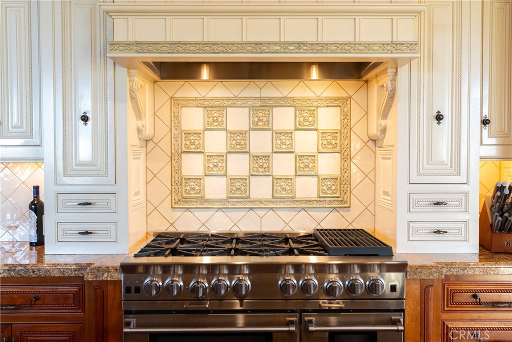 Kitchen range with ornate cream-and-gold backsplash, white cabinetry, and granite counters.