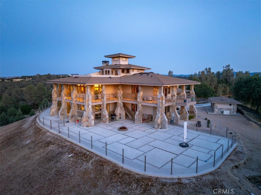 Two-story home with stone supports and wraparound balconies overlooking a broad paved patio at dusk.