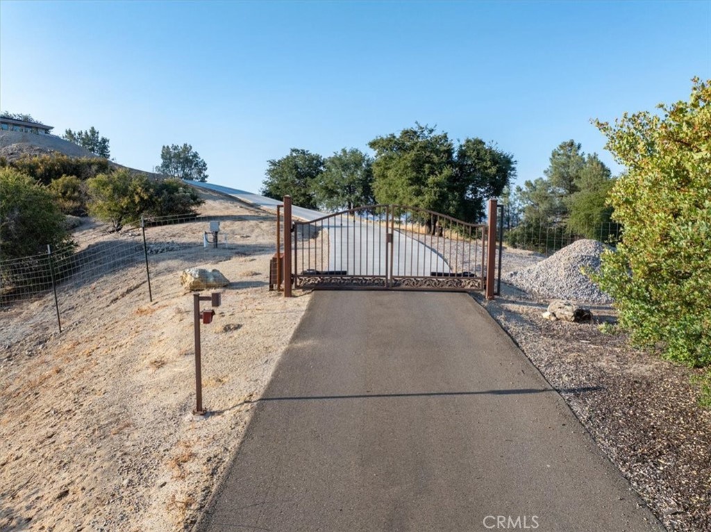 Paved driveway leading to a closed metal gate on a dry, wooded hillside.