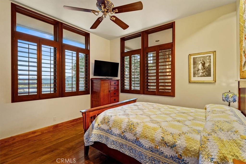 Bedroom with a quilted bed, hardwood floors, and large windows with dark wood shutters.
