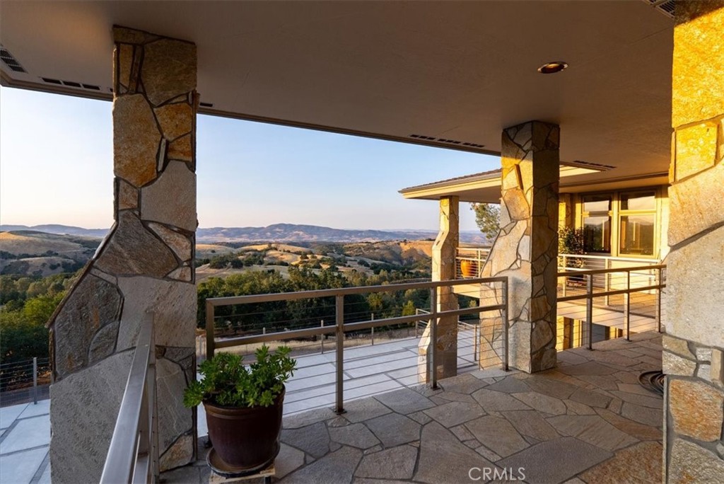 Covered stone patio overlooking rolling, wooded hills, with stone pillars and a metal railing.