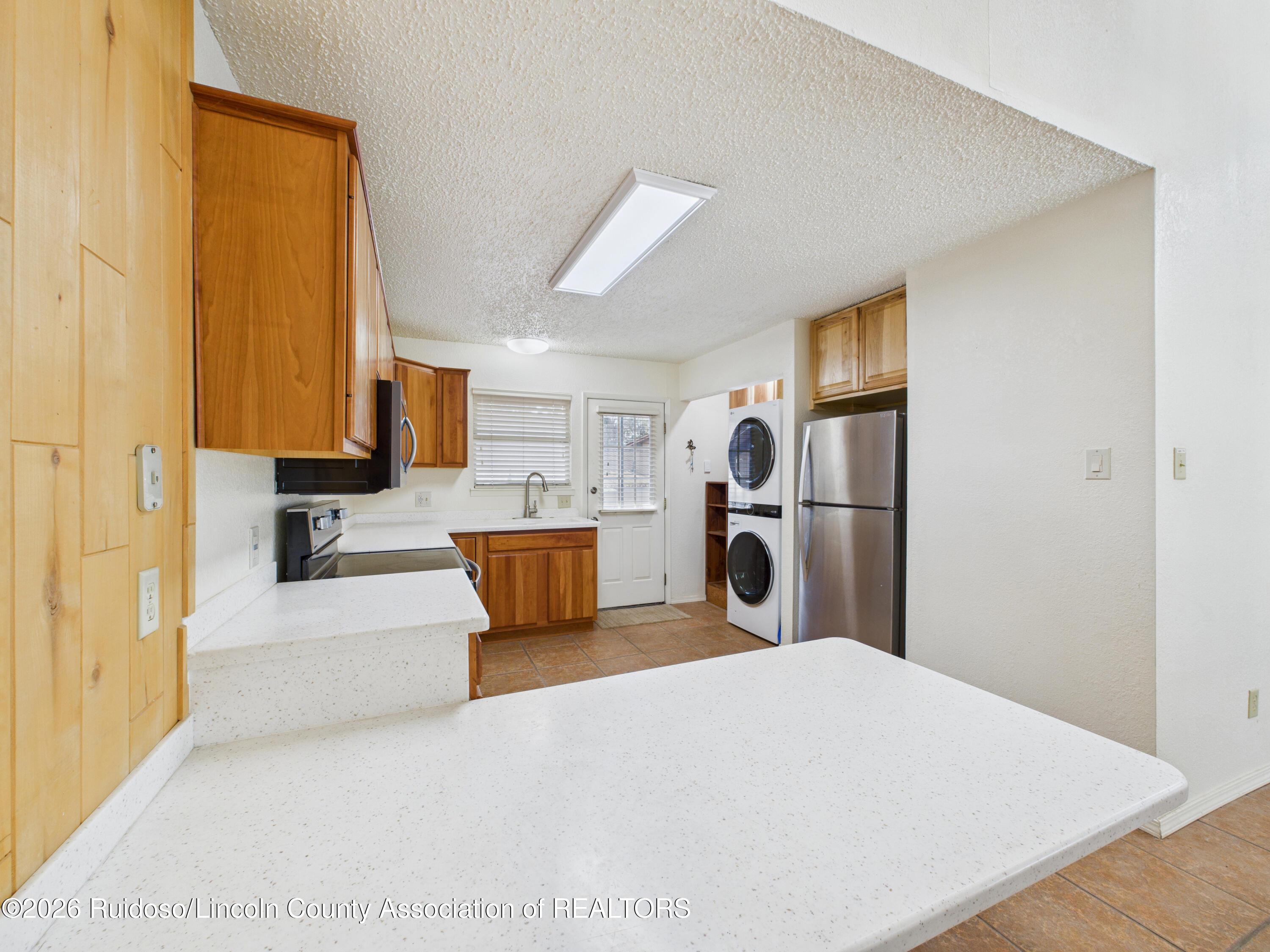 Kitchen with wood cabinets, broad white peninsula, stainless refrigerator, and stacked washer and dryer.